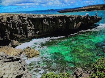 A rugged coastline features dramatic cliffs overlooking a vibrant turquoise sea. Waves crash against the rocks, creating a dynamic and refreshing scene. A person with a sun hat is fishing at the edge of the cliff, while another person sits nearby. The sky is dotted with clouds, adding depth to the expansive view.