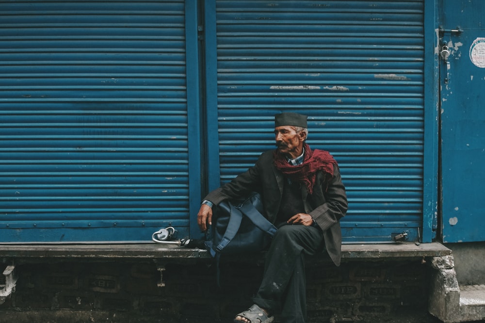 Hombre sentado en un banco gris frente a la puerta de la persiana azul