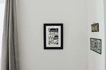 A minimalistic interior wall with a black-framed artwork featuring stylized text. On either side of the artwork are vintage-styled signs, one reading 'BUENOS AIRES' and the other resembling a VW Beetle license plate. To the left, a grey curtain hangs down partially covering a window frame.