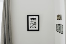 A minimalistic interior wall with a black-framed artwork featuring stylized text. On either side of the artwork are vintage-styled signs, one reading 'BUENOS AIRES' and the other resembling a VW Beetle license plate. To the left, a grey curtain hangs down partially covering a window frame.