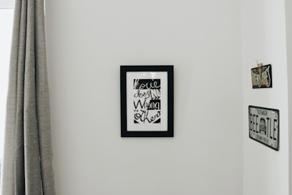 A minimalistic interior wall with a black-framed artwork featuring stylized text. On either side of the artwork are vintage-styled signs, one reading 'BUENOS AIRES' and the other resembling a VW Beetle license plate. To the left, a grey curtain hangs down partially covering a window frame.