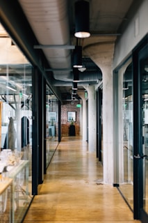 A modern office hallway with polished concrete floors, glass-walled offices on both sides, and exposed industrial elements such as ducts and ceiling pipes. In the distance, a potted plant and framed artwork are visible against a brick wall.