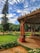 A beautifully landscaped park featuring a large wooden pavilion with intricate carvings and a well-maintained pathway. The pavilion is surrounded by various flowering plants and neatly trimmed grass. In the background, there are tall trees, some people walking, and a partly cloudy blue sky.