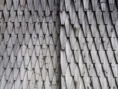 Rows of overlapping wooden shingles create a textured pattern on a surface. The wood appears weathered, showing natural grain and knots. The repetitive geometric design is formed by triangular-shaped shingles with a slightly rough texture.