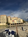 A beachfront condominium with yellow-tinted exteriors and several balconies is situated next to a sandy beach. The sky above is partly cloudy with patches of blue, creating a dramatic backdrop. There are multiple white and blue-striped beach chairs lined up on the sand, with some seagulls visible near the shoreline. Sparse greenery and palm trees are present near the building.