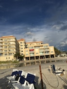A beachfront condominium with yellow-tinted exteriors and several balconies is situated next to a sandy beach. The sky above is partly cloudy with patches of blue, creating a dramatic backdrop. There are multiple white and blue-striped beach chairs lined up on the sand, with some seagulls visible near the shoreline. Sparse greenery and palm trees are present near the building.