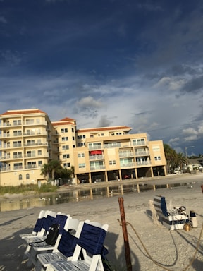 A beachfront condominium with yellow-tinted exteriors and several balconies is situated next to a sandy beach. The sky above is partly cloudy with patches of blue, creating a dramatic backdrop. There are multiple white and blue-striped beach chairs lined up on the sand, with some seagulls visible near the shoreline. Sparse greenery and palm trees are present near the building.