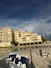 A beachfront condominium with yellow-tinted exteriors and several balconies is situated next to a sandy beach. The sky above is partly cloudy with patches of blue, creating a dramatic backdrop. There are multiple white and blue-striped beach chairs lined up on the sand, with some seagulls visible near the shoreline. Sparse greenery and palm trees are present near the building.