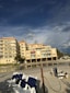 A beachfront condominium with yellow-tinted exteriors and several balconies is situated next to a sandy beach. The sky above is partly cloudy with patches of blue, creating a dramatic backdrop. There are multiple white and blue-striped beach chairs lined up on the sand, with some seagulls visible near the shoreline. Sparse greenery and palm trees are present near the building.