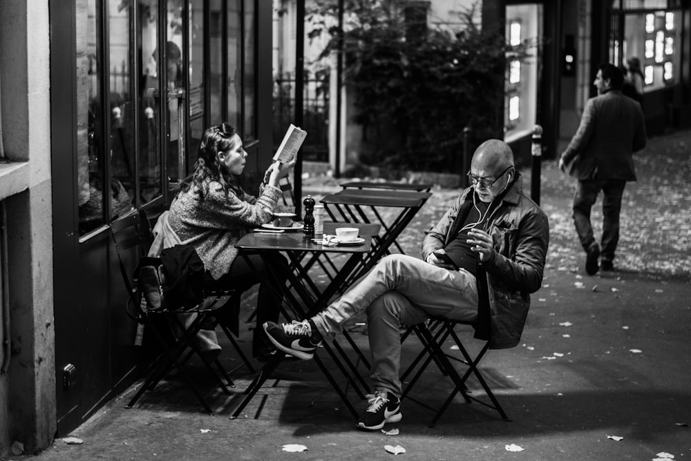 grayscale photo of man and woman sitting beside table holding smartphones