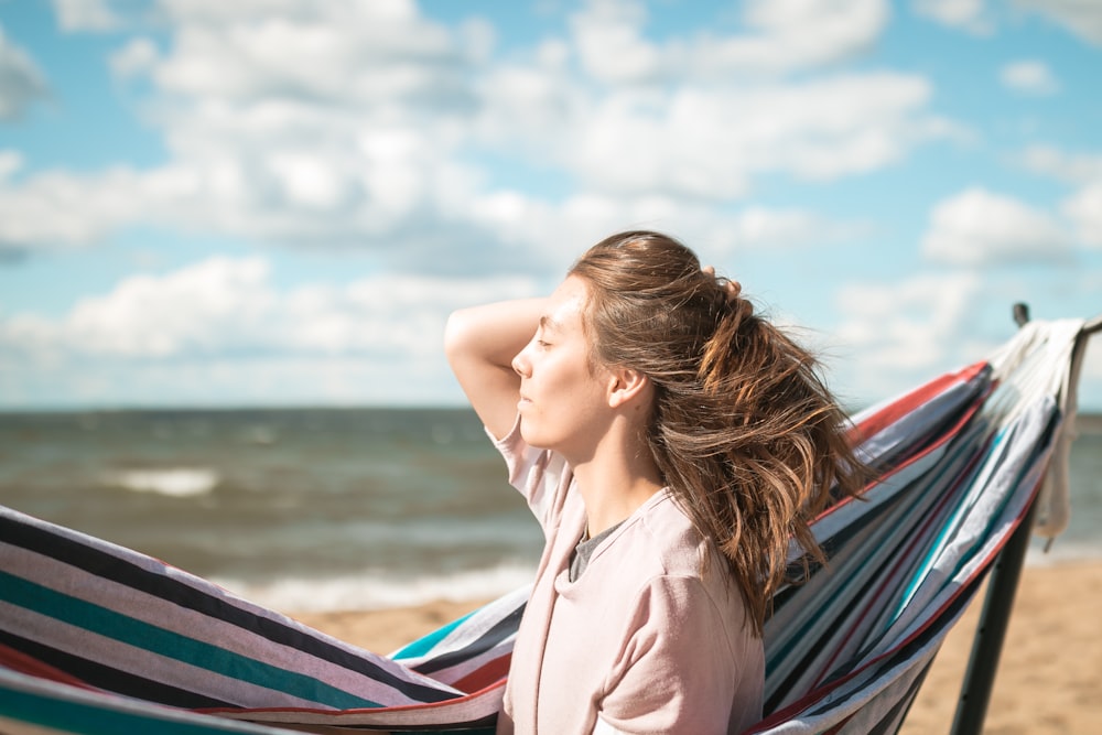 femme tenant ses cheveux en arrière tout en étant assise à côté d’un hamac portable sur le bord de mer pendant la journée
