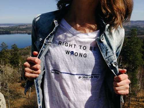 A person in a denim jacket is pulling it open to reveal a white t-shirt with text that reads 'RIGHT TO BE WRONG' and 'be free go your own way.' The background features a scenic view of a forest and a lake under a clear sky.