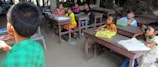 A group of children sit at wooden desks in a classroom setting, appearing attentive and engaged. The classroom has basic furnishings with some books and notebooks on the desks. The atmosphere is rustic, with wooden surroundings and simple decor. One child in a green shirt is turned away, possibly addressing the group.