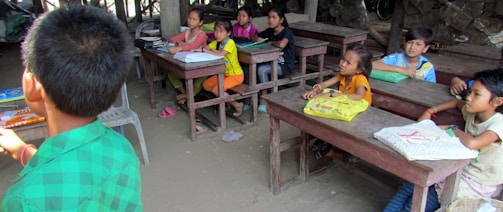 A group of children sit at wooden desks in a classroom setting, appearing attentive and engaged. The classroom has basic furnishings with some books and notebooks on the desks. The atmosphere is rustic, with wooden surroundings and simple decor. One child in a green shirt is turned away, possibly addressing the group.