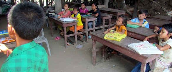 A group of children sit at wooden desks in a classroom setting, appearing attentive and engaged. The classroom has basic furnishings with some books and notebooks on the desks. The atmosphere is rustic, with wooden surroundings and simple decor. One child in a green shirt is turned away, possibly addressing the group.