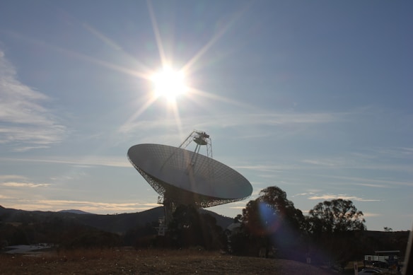 A large satellite dish is positioned against a clear blue sky, with the sun shining brightly near its edge, casting a dramatic glow across the landscape. The surrounding area includes distant hills and scattered trees, creating a sense of openness and vastness.
