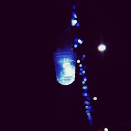 A single illuminated LED light bulb with a soft blue hue is the focal point, set against a dark background. In the distance, a series of blurred blue lights creates a dreamy, bokeh effect.
