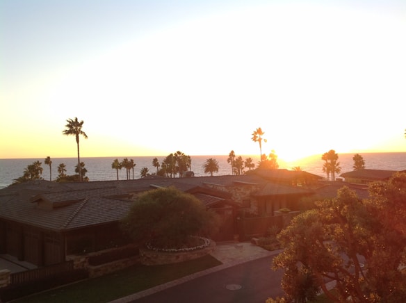 The sun sets over the ocean, casting a warm glow across the sky. Several houses with tiled roofs are nestled among palm trees, and the vibrant colors of the sunset create a serene and picturesque coastal scene.