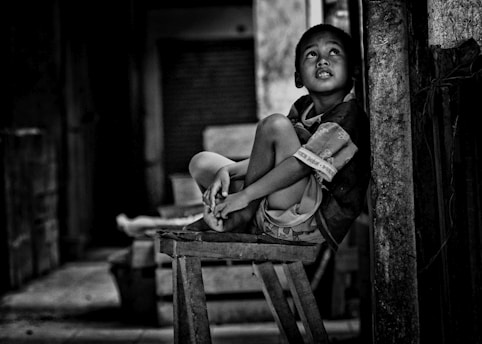 A young boy is sitting on a wooden stool, leaning against a pillar. He looks upwards with a contemplative expression. The scene is monochromatic, highlighting the textures and shadows within a modest, possibly urban interior space.
