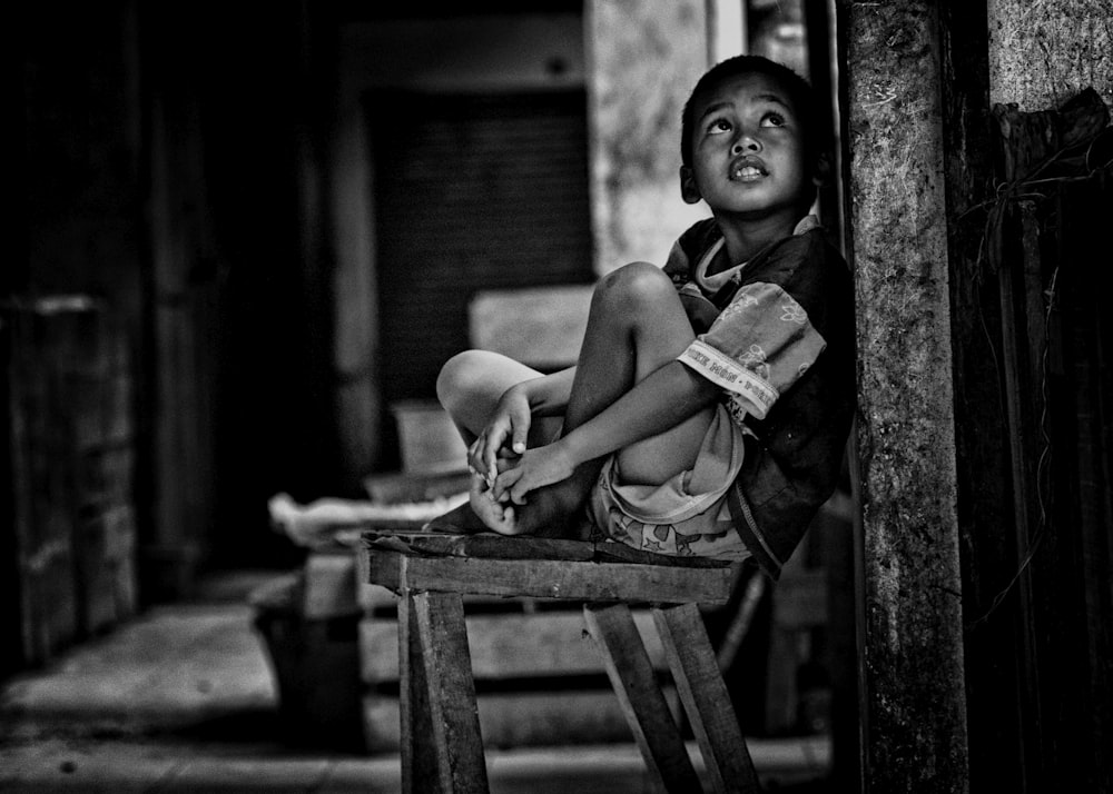 grayscale photo of boy sitting on wooden bench beside wall while looking up