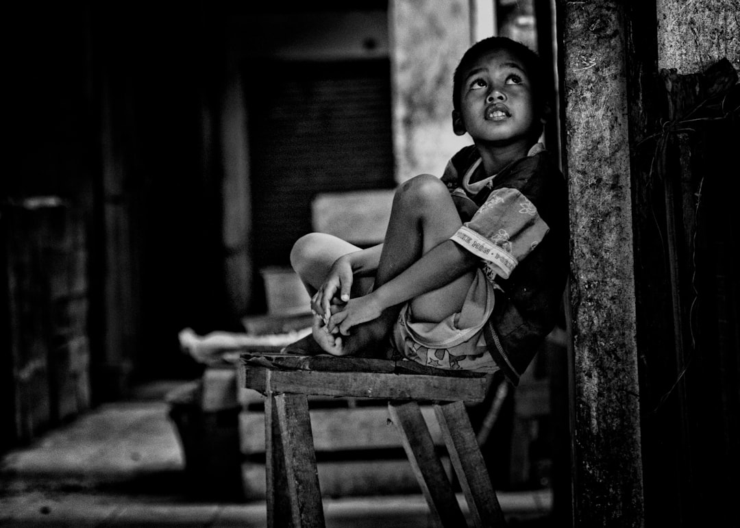 grayscale photo of boy sitting on wooden bench beside wall while looking up