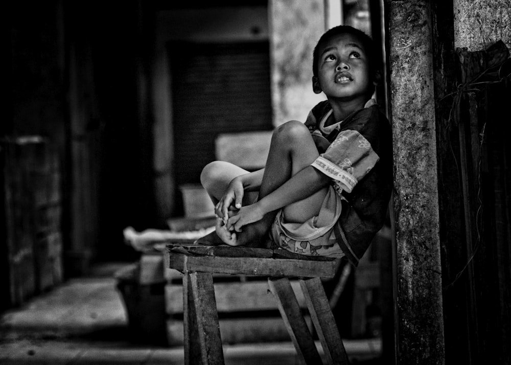 Foto en escala de grises de niño sentado en un banco de madera al lado de la pared mientras mira hacia arriba