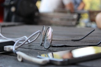 A pair of eyeglasses rests on a wooden table, accompanied by a coiled cable and a smartphone. The table has a textured surface, and the background is softly blurred, suggesting an outdoor setting.