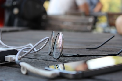 A pair of eyeglasses rests on a wooden table, accompanied by a coiled cable and a smartphone. The table has a textured surface, and the background is softly blurred, suggesting an outdoor setting.