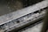 Water droplets are captured mid-air as they fall and splash into a metal gutter filled with water. The surface is reflective with ripples indicating motion, and there is a contrast between the wet, shiny texture of the metal and the droplets.