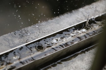 Water droplets are captured mid-air as they fall and splash into a metal gutter filled with water. The surface is reflective with ripples indicating motion, and there is a contrast between the wet, shiny texture of the metal and the droplets.