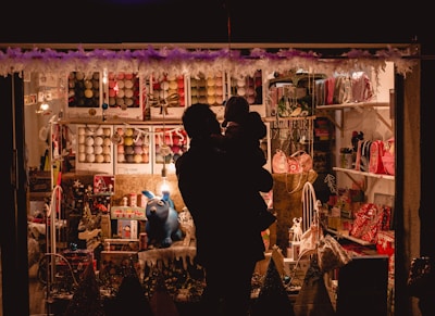 A dimly lit shop window is filled with various colorful items, including toys, bags, and other decorative objects. The window display features a prominent blue rabbit figure. In the foreground, the silhouette of an adult holding a child is visible, facing the window display.