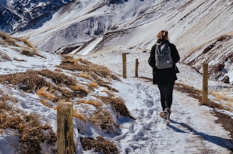 A traveler wearing layered warm clothing walking along a mountain trail with a backpack.