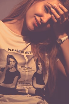 A contemplative woman with long hair rests her head on her hand, wearing a casual t-shirt with monochrome printed images and text. The t-shirt features two identical images of a woman with her hand on her chin. The lighting is warm and soft, casting shadows on her face.