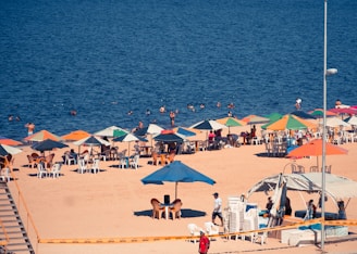 A beach scene with numerous colorful umbrellas and chairs set up on the sand. People are relaxing under the umbrellas, and several others are swimming in the ocean. The water appears deep blue, and the sand is light brown. A lamppost and a canopy are also visible, along with a stack of chairs and equipment near the shoreline.