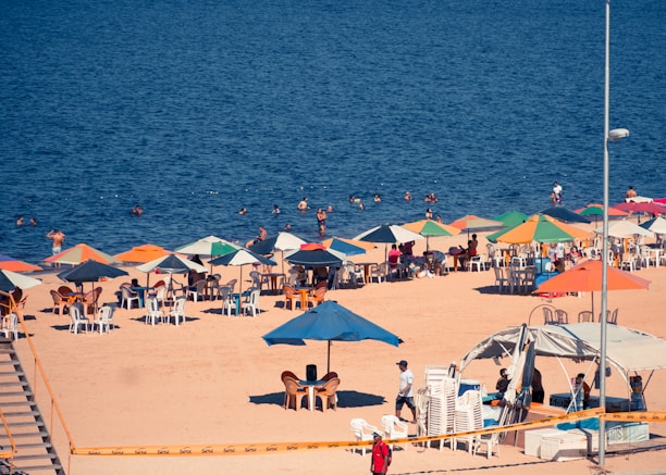 A beach scene with numerous colorful umbrellas and chairs set up on the sand. People are relaxing under the umbrellas, and several others are swimming in the ocean. The water appears deep blue, and the sand is light brown. A lamppost and a canopy are also visible, along with a stack of chairs and equipment near the shoreline.