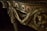Intricately carved wooden detail of furniture featuring ornamental floral patterns and a sculpted animal figure. The design includes flowing drapery motifs and twisted column elements.