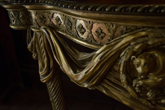 Intricately carved wooden detail of furniture featuring ornamental floral patterns and a sculpted animal figure. The design includes flowing drapery motifs and twisted column elements.