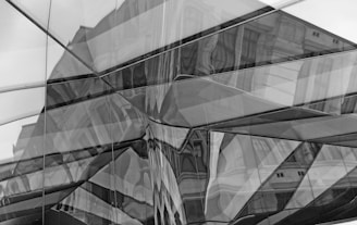 A complex reflection of a building in a series of overlapping, angular glass panes. The architecture appears distorted due to the refractions and geometry of the glass surfaces, creating an abstract and intricate visual effect.