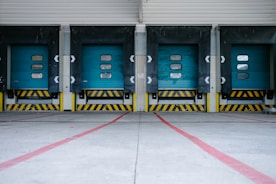 Five industrial loading dock doors painted in teal, lined side by side. Each door features small rectangular windows and is framed by black and yellow warning stripes. The concrete ground in front of the doors is marked with red lines.