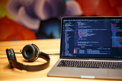 A laptop displaying programming code on the screen sits on a wooden table. Next to it, a pair of black headphones rests on the table surface. The background features a colorful, abstract mural with soft, blurred shapes.