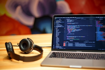 A laptop displaying programming code on the screen sits on a wooden table. Next to it, a pair of black headphones rests on the table surface. The background features a colorful, abstract mural with soft, blurred shapes.