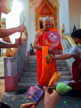 A monk in orange robes is standing on steps leading up to a temple adorned with ornate carvings. Several people around the monk are participating in a ceremonial activity, offering items such as banknotes and flowers. The sunlight is casting a bright glow onto the scene, highlighting the cultural and spiritual setting.