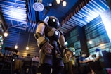A humanoid robot with glowing lights stands in a dimly lit restaurant interior. The ceiling features industrial-style lighting and exposed beams, creating a modern atmosphere. People are seated at tables in the background, engaged in conversation.