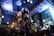 A humanoid robot with glowing lights stands in a dimly lit restaurant interior. The ceiling features industrial-style lighting and exposed beams, creating a modern atmosphere. People are seated at tables in the background, engaged in conversation.