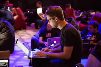 A group of people are seated indoors, focused intently on working with laptops. The environment is informal and relaxed, with individuals sitting on beanbags and surrounded by dim lighting. Most people appear absorbed in their screens, suggesting a collaborative or hackathon-style event.