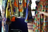 Colorful, patterned garments hang in a market stall. The fabrics feature intricate, vibrant geometric designs in a mix of yellow, blue, orange, and other colors. A hand points to a skirt with an elaborate circular pattern, while woven baskets are visible below.