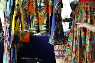 Colorful, patterned garments hang in a market stall. The fabrics feature intricate, vibrant geometric designs in a mix of yellow, blue, orange, and other colors. A hand points to a skirt with an elaborate circular pattern, while woven baskets are visible below.