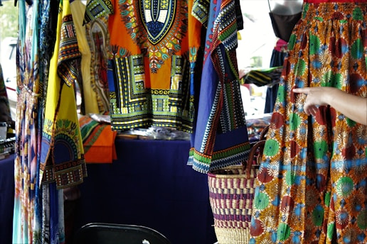 Colorful, patterned garments hang in a market stall. The fabrics feature intricate, vibrant geometric designs in a mix of yellow, blue, orange, and other colors. A hand points to a skirt with an elaborate circular pattern, while woven baskets are visible below.