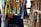 Colorful, patterned garments hang in a market stall. The fabrics feature intricate, vibrant geometric designs in a mix of yellow, blue, orange, and other colors. A hand points to a skirt with an elaborate circular pattern, while woven baskets are visible below.