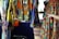 Colorful, patterned garments hang in a market stall. The fabrics feature intricate, vibrant geometric designs in a mix of yellow, blue, orange, and other colors. A hand points to a skirt with an elaborate circular pattern, while woven baskets are visible below.
